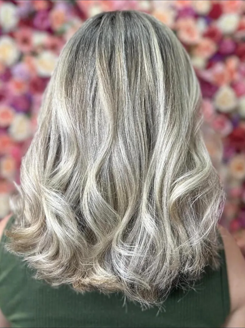 Cabelo com mechas claras sobre base acinzentada, ondulado, visto de costas diante de um painel de rosas