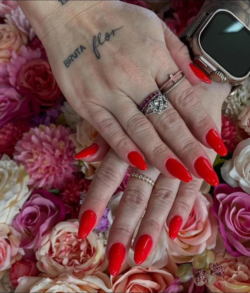 Mãos com unhas em gel vermelhas, amendoadas, sobre um painel de rosas